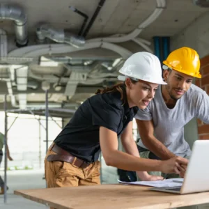 Two construction workers reviewing plans on a laptop at a site.